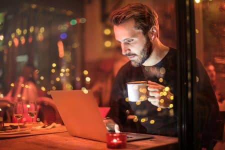 Comment choisir les meilleurs outils de capture d’écran gratuits A young man with a beard is working on a laptop at a cozy cafe with warm lighting, holding a coffee cup.