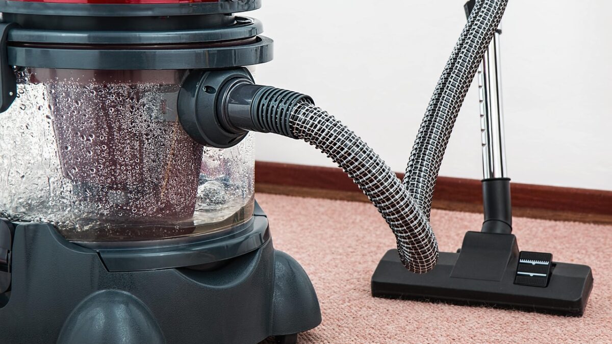 Aspirateur à cuve transparente rouge et gris posé sur une moquette rose, avec un long tuyau flexible noir relié à une brosse de sol noire.