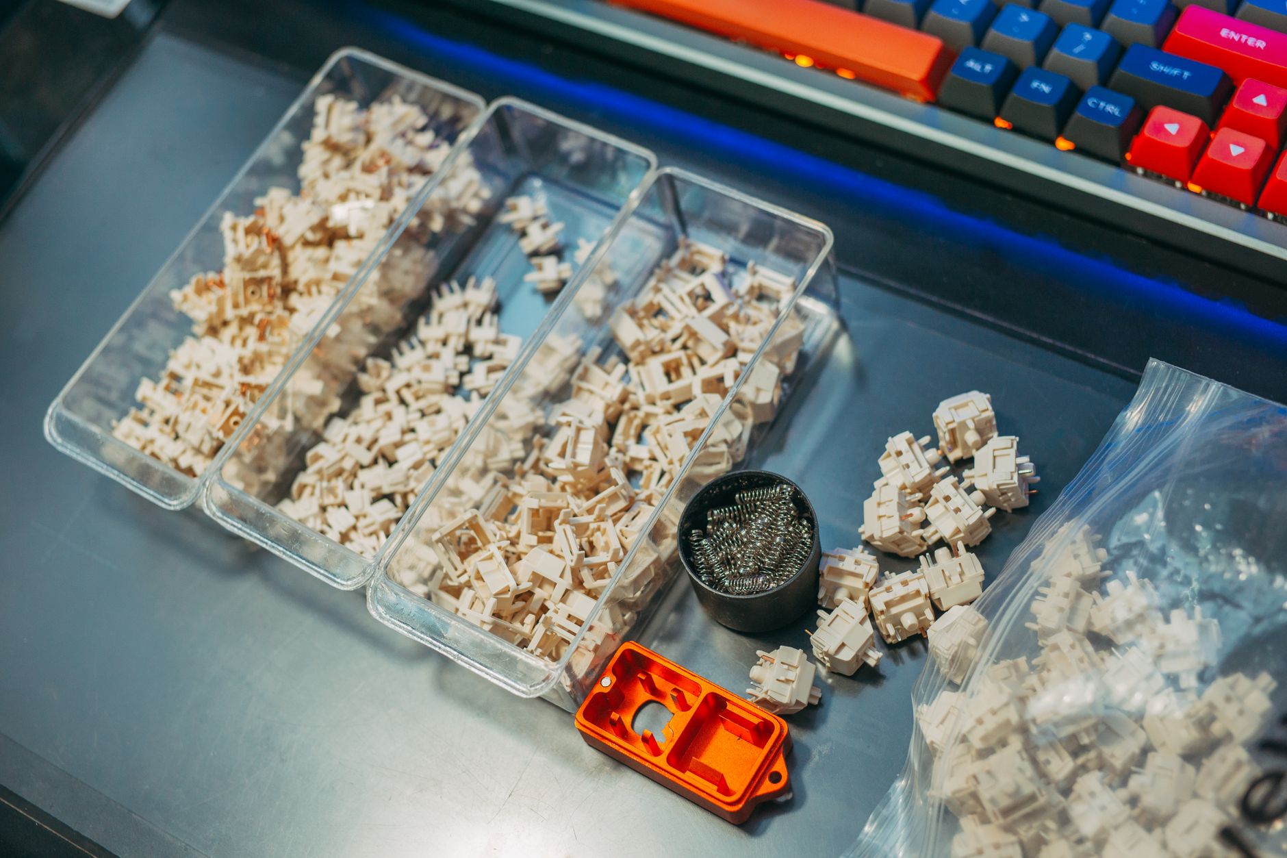 Interrupteurs de clavier mécanique beiges dans des bacs transparents, avec ressorts, petit outil orange et clavier coloré en haut de l’image