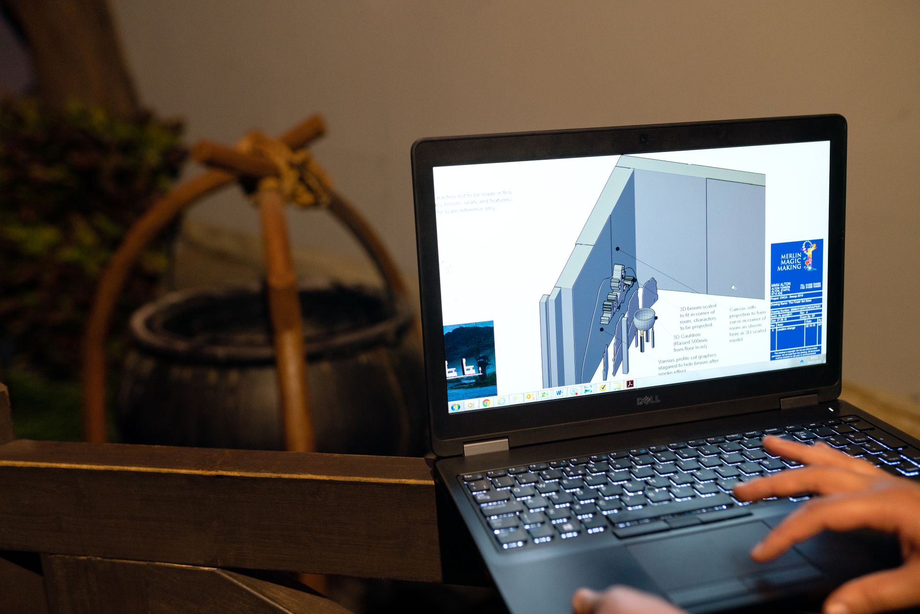 Mains sur le clavier d’un ordinateur portable Dell dont l’écran affiche un schéma technique en 3D, devant un chaudron en arrière-plan flou.