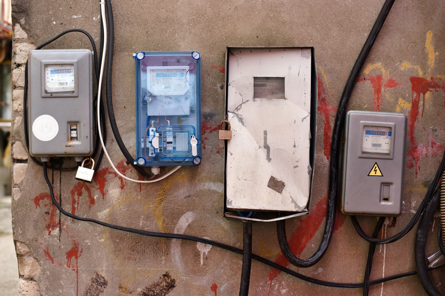 Quatre compteurs électriques fixés sur un mur dégradé, avec des câbles noirs apparents, des cadenas et des traces de peinture rouge.