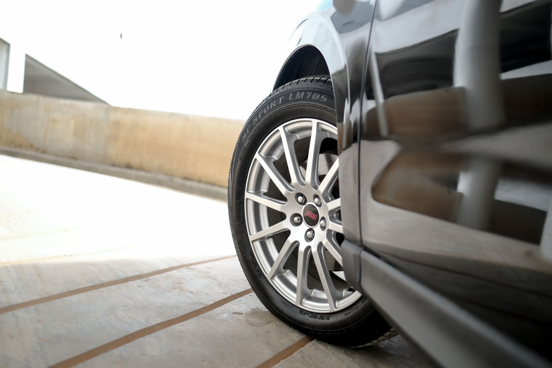 Roue en alliage argenté à rayons fins et pneu noir sur le côté d’une voiture sombre, garée sur une rampe en béton.
