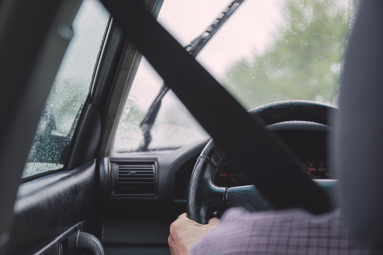 Un conducteur ceinturé tient le volant sous la pluie, vu depuis l’arrière de l’habitacle