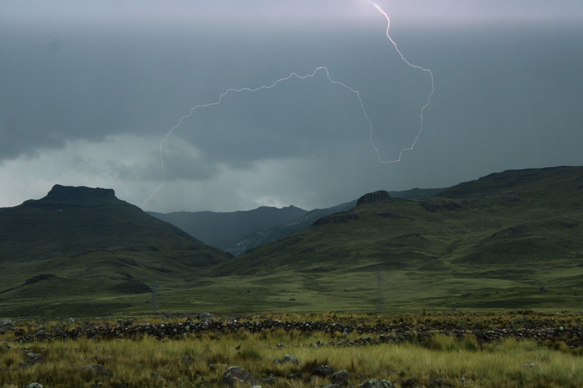 Un éclair zèbre un ciel d’orage au-dessus de collines herbeuses, avec un muret de pierres et des herbes hautes au premier plan.