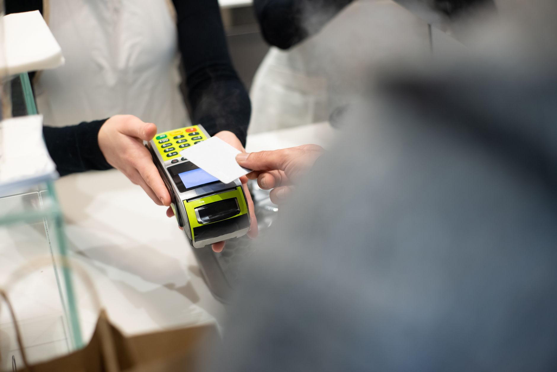 Une personne approche une carte blanche d’un terminal de paiement jaune et vert tenu par une vendeuse au comptoir d’un commerce.