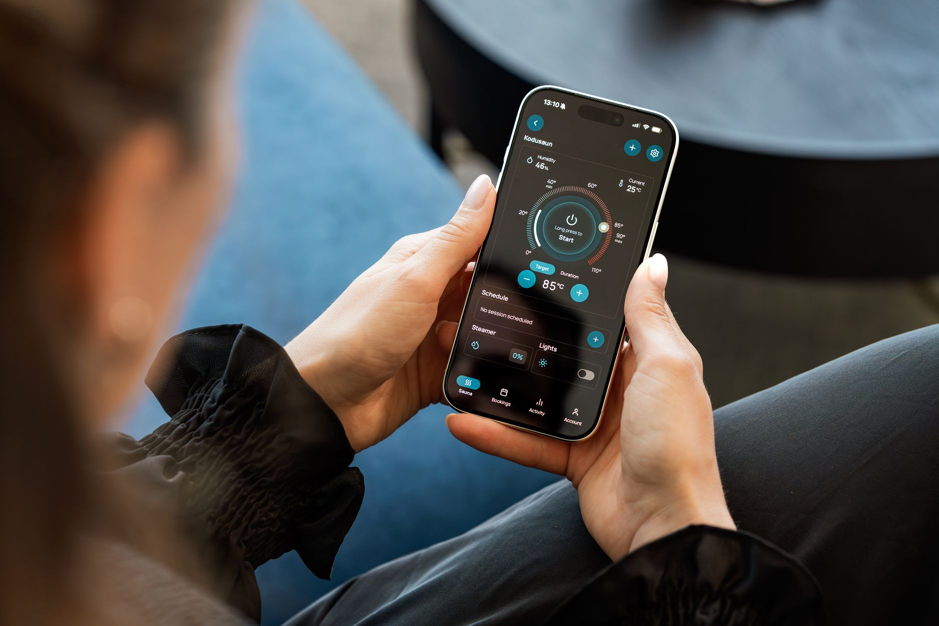 Une personne tient un smartphone affichant une application de commande de sauna connecté, assise près d’un canapé bleu.